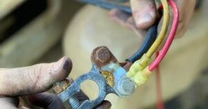 Hands holding a corroded battery terminal with multi-colored wires in a repair setting.